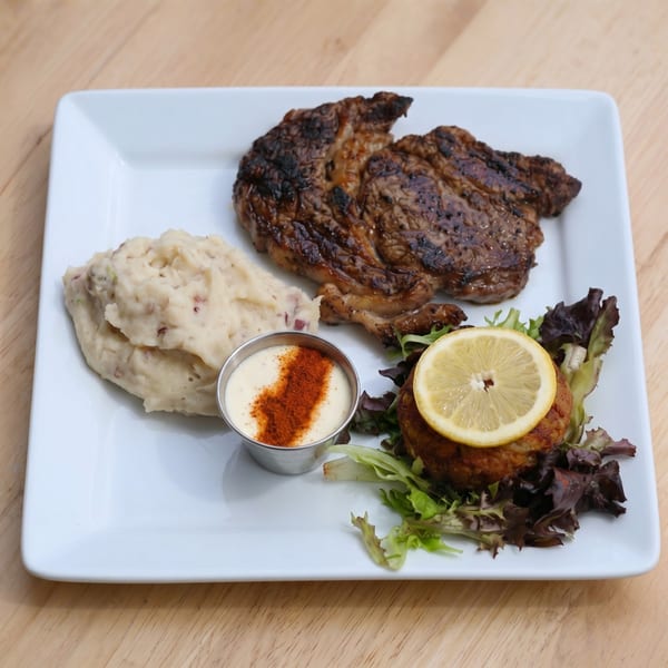 Grilled steak, mashed potatoes, and a crab cake with lemon and sauce on a white plate.