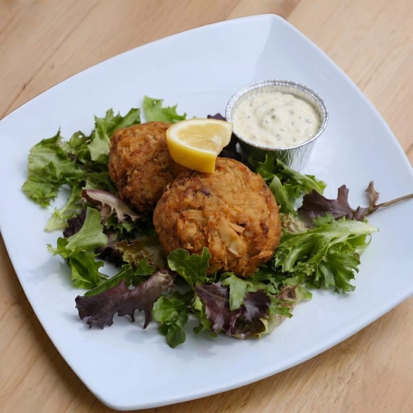 Two golden-brown crab cakes on a bed of mixed greens, with a lemon wedge and tartar sauce.